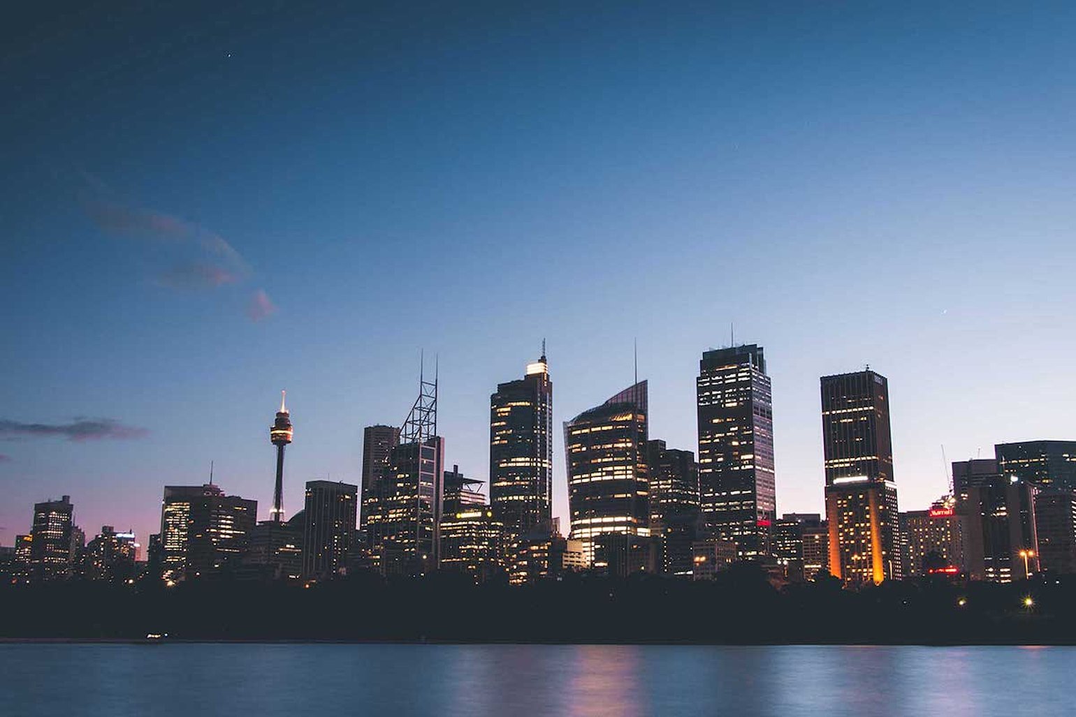 Skyline einer Stadt bei Daemmerung mit beleuchteten Hochhaeusern und reflektierendem Wasser im Vordergrund